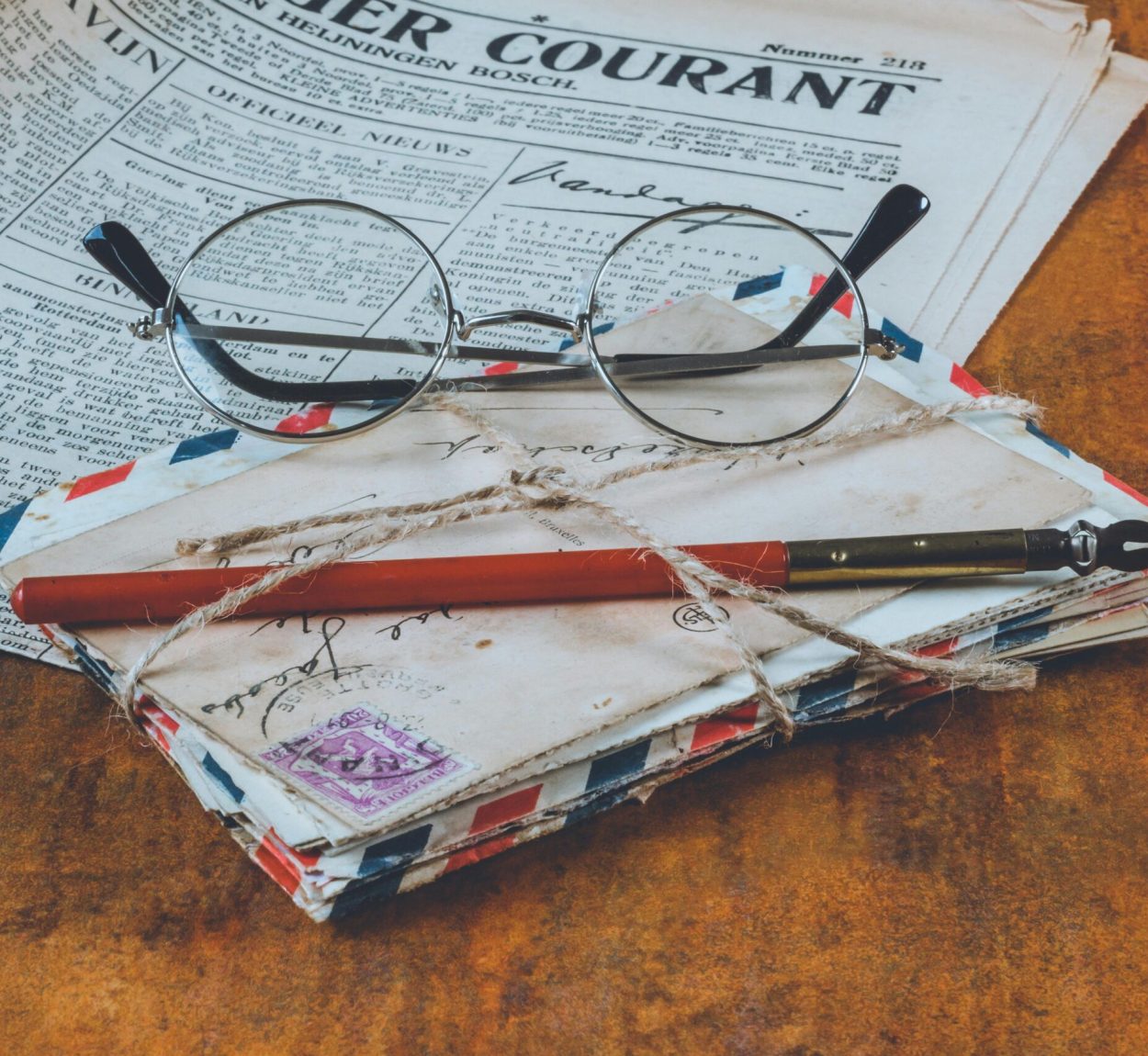Historical letters and a fountain pen on a vintage wooden table, alongside eyeglasses and a newspaper.